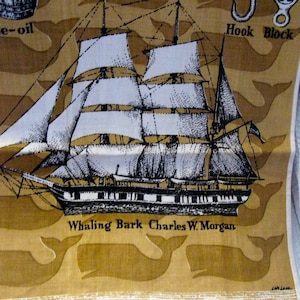 May include: A tan fabric tea towel featuring a black and white illustration of the whaling bark Charles W. Morgan. The design includes repeating whale silhouettes in the background and text labels reading Whale-oil, Hook, Block, and Whaling Bark Charles W. Morgan.