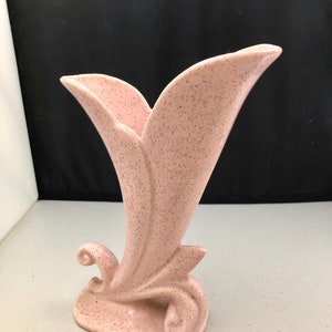 May include: A pink speckled ceramic vase with a unique, stylized design. The vase has a wide opening and a narrow base, with a curved, leaf-like design.