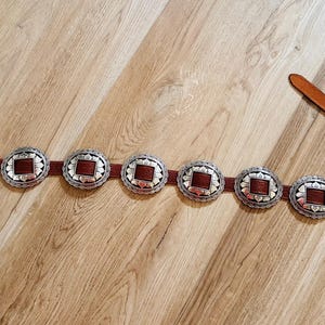 May include: A brown leather belt with seven silver-toned, circular buckle accents. Each buckle has a square, reddish-brown center. The belt is laid out on a light brown wooden surface.