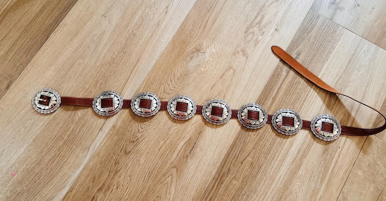 May include: A brown leather belt with seven silver-toned, circular buckle accents. Each buckle has a square, reddish-brown center. The belt is laid out on a light brown wooden surface.