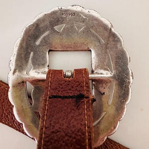 May include: A silver-colored, scalloped-edge belt buckle with a rectangular opening and a brown leather strap. The buckle has a decorative design with cut-out shapes. The leather strap is stitched to the buckle.
