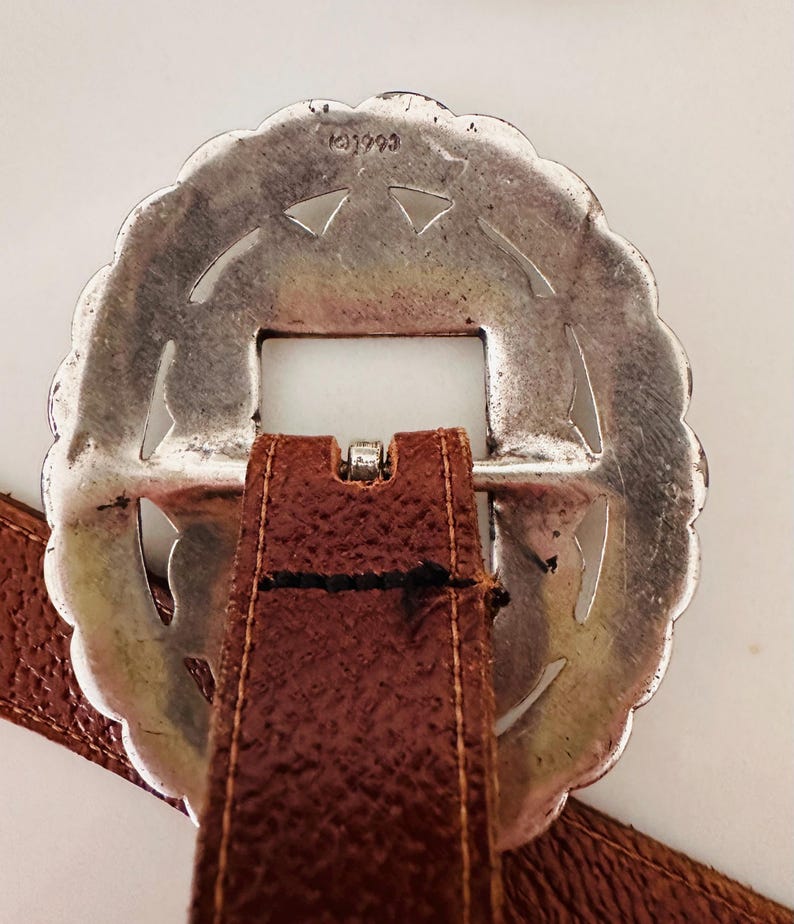 May include: A silver-colored, scalloped-edge belt buckle with a rectangular opening and a brown leather strap. The buckle has a decorative design with cut-out shapes. The leather strap is stitched to the buckle.
