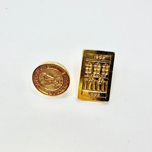 May include: Two gold-colored pins. One pin features a building with the text "1952" and "1992". The other pin features a circular design with the text "Givenchy" and "Fortissimo".