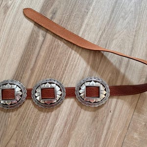 May include: A brown leather belt with three silver-colored, scalloped-edge buckles. Each buckle has a square, brown leather center. The belt is displayed on a light brown wooden surface.