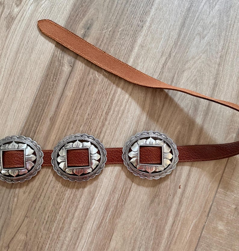 May include: A brown leather belt with three silver-colored, scalloped-edge buckles. Each buckle has a square, brown leather center. The belt is displayed on a light brown wooden surface.