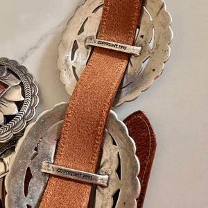 May include: Close-up of a brown leather belt with ornate silver-toned buckles. The buckles have a decorative filigree design. The text "COPYRIGHT 1993" is visible on the metal hardware.