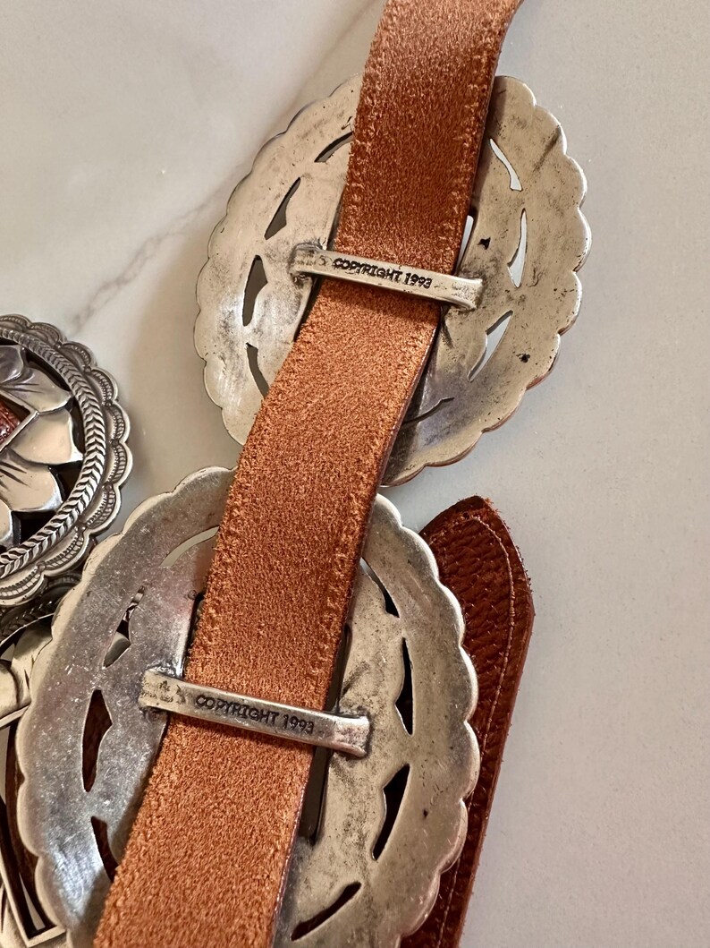 May include: Close-up of a brown leather belt with ornate silver-toned buckles. The buckles have a decorative filigree design. The text "COPYRIGHT 1993" is visible on the metal hardware.