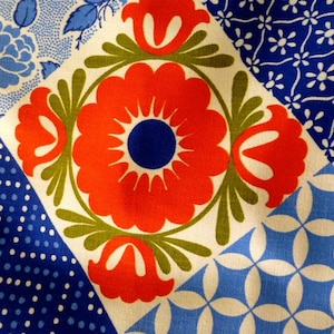 May include: Close-up of a fabric with a patchwork design. The design features a large red flower with a blue center, surrounded by green leaves. Other squares have blue floral and polka dot patterns on a white background.