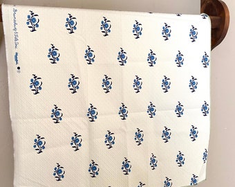 May include: A rectangular piece of off-white fabric featuring a repeating pattern of small blue floral sprigs. The text "Brunschwig & Fils Inc." is printed along the left edge. The fabric has a textured surface and a finished hem along the bottom edge.