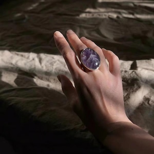 May include: A ring featuring a large, oval-shaped purple amethyst gemstone set in a polished silver band. The stone displays natural white veining and a smooth, rounded surface, securely held by a simple metal bezel setting on the finger.