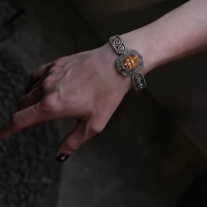May include: A silver-toned metal cuff bracelet featuring an oval-shaped amber-coloured stone at the centre. The bracelet band is decorated with intricate swirling metal patterns on both sides of the central stone setting, creating a detailed look.