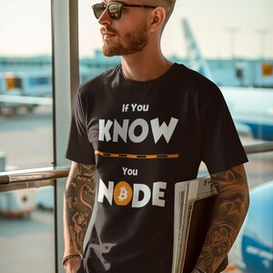 May include: Black t-shirt with the text "If You Know You Node" in white, with a Bitcoin symbol. The shirt is worn by a person wearing sunglasses and standing near a window. The background includes an airplane.