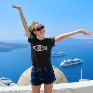 May include: A black t-shirt with a Bitcoin symbol inside a fish outline. The person is wearing sunglasses and denim shorts, with arms raised against a backdrop of a blue sea and sky.