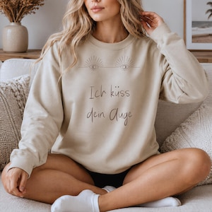 Könnte beinhalten: Ein beigefarbenes Sweatshirt mit Rundhalsausschnitt und einer dunkelbraunen Grafik von zwei stilisierten Augen mit strahlenförmigen Linien über dem Text Ich küss dein Auge. Das langärmelige Kleidungsstück hat gerippte Bündchen am Kragen.