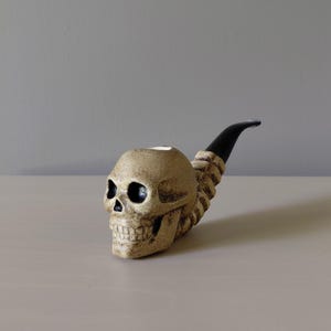 May include: A tobacco pipe featuring a beige, textured skull-shaped bowl with deep eye sockets and a nasal cavity. The stem of the pipe is black, smooth, and curved, extending from the back of the skull, which is sculpted to resemble a spinal column.