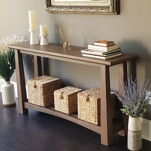 Puede incluir: Una consola de madera marrón con una balda inferior que contiene tres cestas de almacenamiento tejidas. La mesa exhibe velas, libros y un difusor de caña. Un espejo decorativo cuelga sobre la mesa, y jarrones con plantas a los lados.