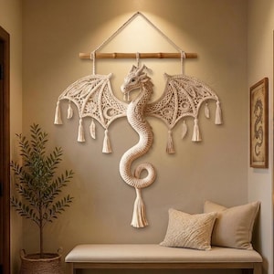 Può includere: Un arazzo da parete a forma di drago in macramè color crema. Il drago ha ali dettagliate, un corpo lungo e curvo e un nappa all'estremità. È sospeso da un tassello di legno con una corda.