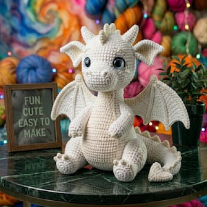 Peut inclure: Un dragon en crochet blanc cass&eacute; fait &agrave; la main, avec de grands yeux bleus, des ailes et une queue &eacute;pineuse. Le dragon est assis sur une surface en marbre vert. Une petite pancarte encadr&eacute;e indique "FUN CUTE EASY TO MAKE". De la laine color&eacute;e et une plante en pot sont en arri&egrave;re-plan.