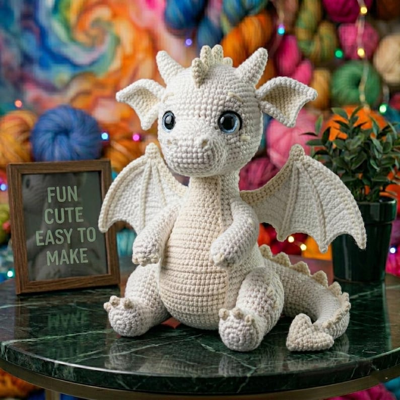 Peut inclure: Un dragon en crochet blanc cass&eacute; fait &agrave; la main, avec de grands yeux bleus, des ailes et une queue &eacute;pineuse. Le dragon est assis sur une surface en marbre vert. Une petite pancarte encadr&eacute;e indique "FUN CUTE EASY TO MAKE". De la laine color&eacute;e et une plante en pot sont en arri&egrave;re-plan.