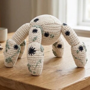 May include: A handmade, crocheted spider toy in cream color with black star-shaped accents. The spider has six legs with green vine-like details. The toy is sitting on a wooden table.