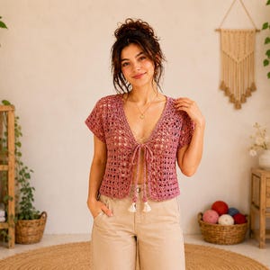 May include: A short-sleeved, open-front, crochet top in a dusty rose color. The top features a V-neckline with a tie closure and small tassels. The model is wearing beige pants. The background includes a wooden shelf, plants, and a woven wall hanging.