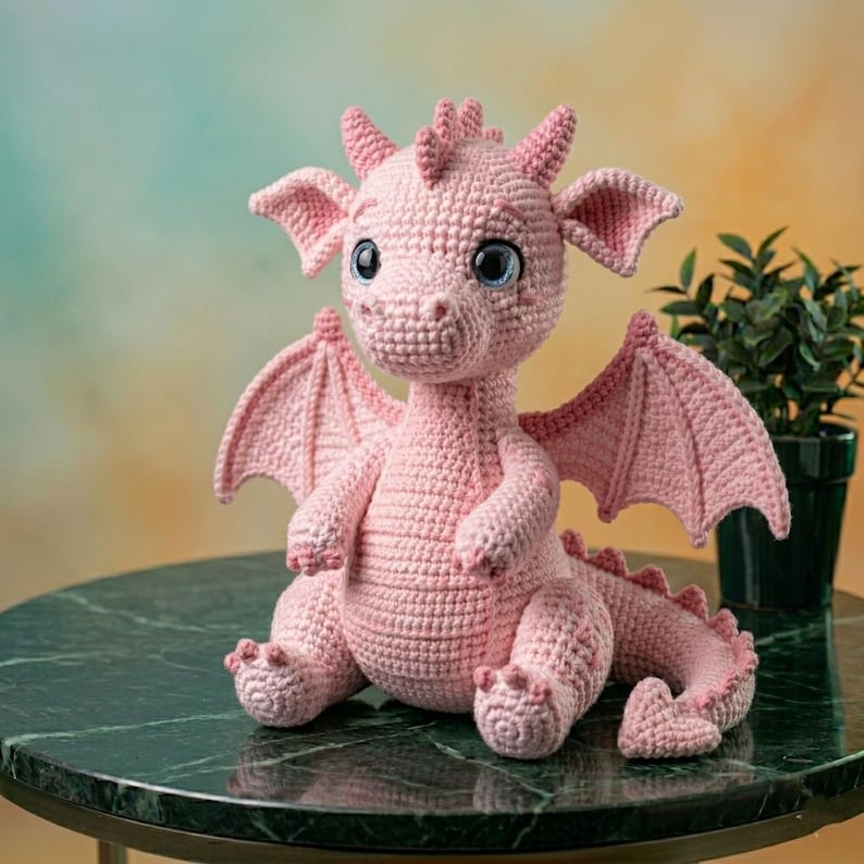 Peut inclure: Un dragon rose en crochet fait &agrave; la main avec des yeux bleus, des ailes et des cornes. Le dragon est assis sur une table en marbre vert. Le dragon est un jouet en peluche.