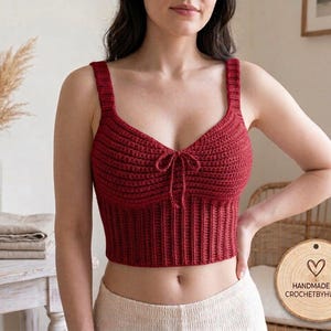 May include: A deep red crochet crop top featuring a sweetheart neckline, thick shoulder straps, and a ribbed hem. The centre of the bodice includes a small decorative tie. A circular wooden tag displays the text HANDMADE BY CROCHETBYHUNZA.