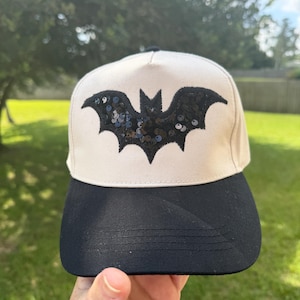 May include: A baseball cap featuring a cream-colored front panel with a black sequined bat emblem centered on the front. The cap has a solid black brim and a black button at the top, constructed with a structured crown and a curved visor design.