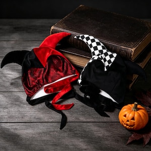 May include: Two jester hats with pointed ears and tie closures. One hat is made of black and red patterned velvet fabric. The second hat features a black and white checkered pattern on one side and solid black fabric on the other side.