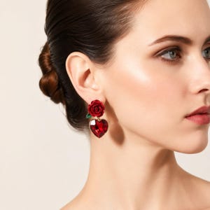 May include: A drop earring featuring a deep red rose flower at the top, connected to a faceted heart-shaped red crystal pendant. The rose includes a small green leaf detail at the base, and the entire piece is suspended from a gold-toned metal setting.