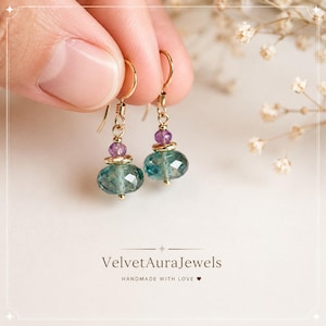 Könnte beinhalten: Ein Paar Ohrhänger mit facettierten blaugrünen Perlen, kleinen violetten Akzentperlen und goldfarbenen Metallelementen. Die Ohrringe hängen an goldfarbenen Haken. Der Text VelvetAuraJewels HANDMADE WITH LOVE ist unten aufgedruckt.