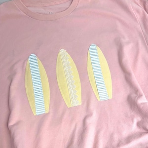 May include: A light pink t-shirt featuring three yellow, oval-shaped fabric appliqués arranged horizontally. The two outer appliqués are decorated with vertical light blue and white striped ribbon, while the centre one features white lace trim.