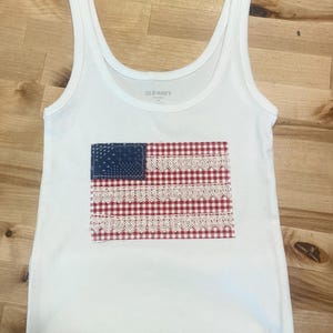May include: White tank top with a patriotic American flag design. The flag features a blue square with white stars, red and white gingham stripes, and white lace trim. The tank top is made of a ribbed material.