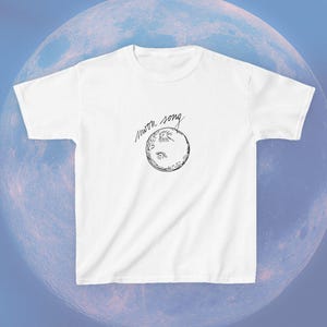 May include: A white short-sleeved t-shirt featuring a black line-art illustration of a moon centred on the chest. Above the moon, the text "moon song" is written in a cursive, handwritten script style. The shirt has a crew neckline.