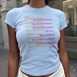 Könnte beinhalten: Ein hellblaues T-Shirt mit kurzen Ärmeln und aufgedrucktem Liedtext in orangefarbener und pinker Schrift. Der Text lautet: Alright, Hey, I think you're perfect, You say that to everyone, 'Kay, well, is it working?, Yeah, but don't tell anyone, Ah, you seem uncertain, Well, you're just having fun, Oh, you don't know me, Mm-hmm, Alright then, honey, What is it this time?, Well, see, I got money, Oh, you think you're big time?, No, just being funny, Oh, you're Mr. Big Light?, Ah, you don't know me, Uh-huh.