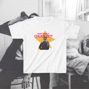 以下が含まれることがあります： 胸元に黄色い星のグラフィックがプリントされた白い半袖Tシャツ。星の中にはサングラスをかけた人物のモノクロ写真があり、その上にピンク色の文字でWHAT WOULD CHUCK DO?と書かれています。