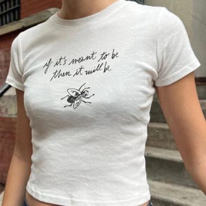 May include: White short-sleeved t-shirt with the text "if it's meant to be then it will be" in black script. Below the text is a black and white illustration of a fly. The shirt is a fitted style.