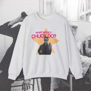 Op de afbeelding: Een lichtgrijs sweatshirt met ronde hals en een grote oranje stergrafiek op de borst. In het midden van de ster staat een zwart-witfoto van een persoon met een zonnebril die een klein voorwerp vasthoudt, met de tekst WHAT WOULD CHUCK DO?