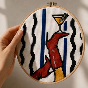 May include: A circular wooden embroidery hoop containing a cross-stitch design on white fabric. The pattern features a red boot holding a yellow cocktail glass with olives, set against a background of vertical black wavy lines and blue stripes.