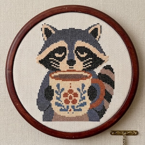 May include: A circular wooden embroidery hoop frames a cross-stitch piece featuring a gray and black raccoon holding a cream-colored mug. The mug is decorated with a small red flower and blue leaves. The raccoon has a striped tail and closed eyes.