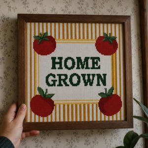 May include: A square cross-stitch embroidery in a dark wood frame featuring the text HOME GROWN in dark green letters. The design includes four red tomatoes with green stems, one in each corner, set against a background of vertical yellow stripes.