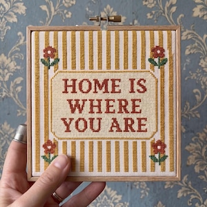 May include: A square cross-stitch embroidery in a light wood frame featuring vertical yellow and white stripes. The center contains the text "HOME IS WHERE YOU ARE" in rust-colored thread, surrounded by four small red flowers with green stems.
