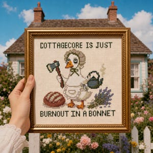 May include: A framed cross-stitch embroidery featuring a white duck wearing a bonnet and apron, holding an axe and a steaming teapot. Beside the duck is a loaf of bread and lavender. The text reads: COTTAGECORE IS JUST BURNOUT IN A BONNET.