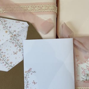 May include: A white stationery card and matching envelope featuring a delicate floral, butterfly, and bird illustration in soft pink and muted tones. The set is displayed alongside gift-wrapped packages adorned with sheer pink ribbons and lace.