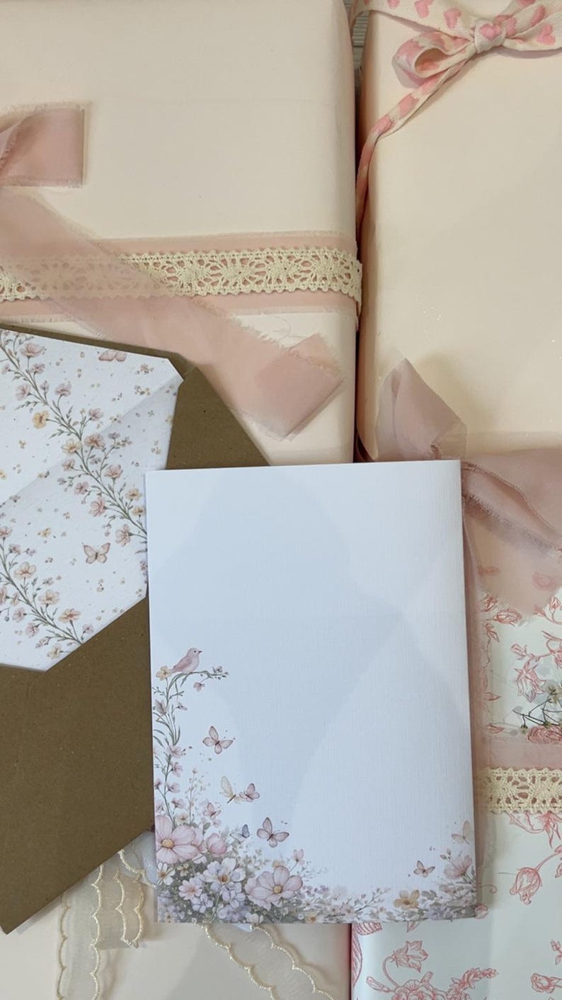 May include: A white stationery card and matching envelope featuring a delicate floral, butterfly, and bird illustration in soft pink and muted tones. The set is displayed alongside gift-wrapped packages adorned with sheer pink ribbons and lace.