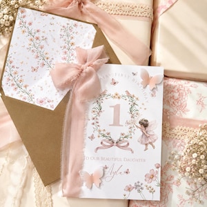 May include: A fairy-themed first birthday card with a floral wreath, butterflies, and the number 1. The card is decorated with a pink ribbon and is accompanied by a matching envelope and decorative gift boxes.
