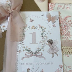 May include: A white rectangular greeting card featuring the text Fairy First Birthday and Our Beautiful Daughter with the name Ayla. The card displays a floral wreath, a number one, a fairy illustration, and three-dimensional butterfly accents.