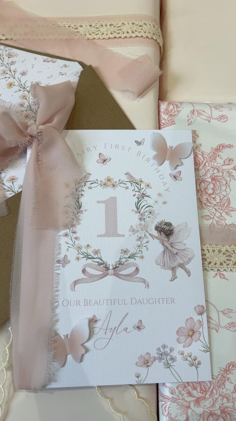 May include: A white rectangular greeting card featuring the text Fairy First Birthday and Our Beautiful Daughter with the name Ayla. The card displays a floral wreath, a number one, a fairy illustration, and three-dimensional butterfly accents.