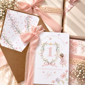 May include: A white birthday card with the text Happy Fairy First Birthday and To Our Beautiful Daughter Ayla, featuring a large number one, a floral wreath, and a fairy illustration. The card is tied with a pink ribbon and paired with a floral envelope.