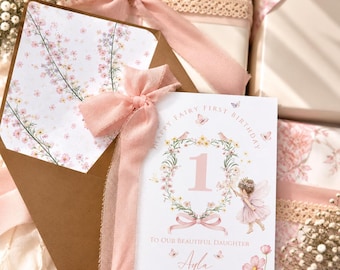 Puede incluir: Una tarjeta de cumpleaños blanca con el texto Happy Fairy First Birthday y To Our Beautiful Daughter Ayla, que presenta un número uno grande, una corona floral y una ilustración de hada. La tarjeta lleva un lazo rosa y un sobre con flores.